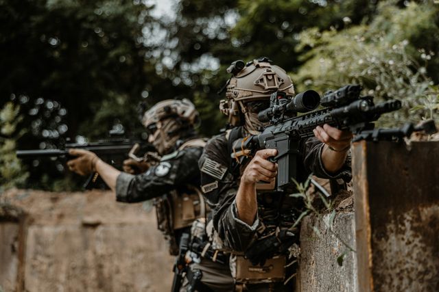 Plusieurs joueurs d'airsoft d'une même équipe tirent depuis un muret en ruine, en tenue camouflée