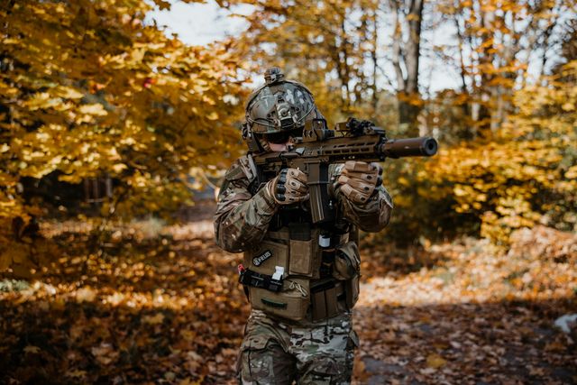 Joueur d'airsoft équipé, réplique épaulée, au milieu d'un sous-bois aux feuilles d'automne