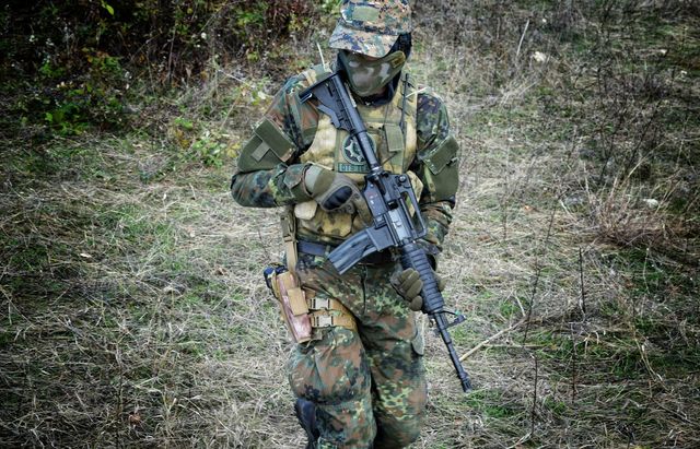 Joueur d'airsoft en tenue camouflée et gilet tactique, réplique en main, debout dans les hautes herbes