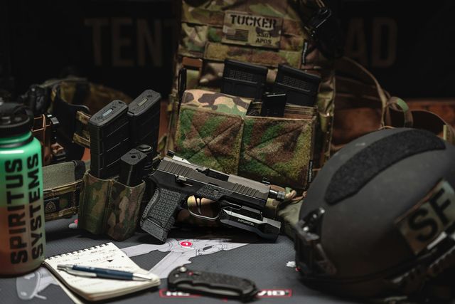 Setup airsoft exposé sur table : pistolet, plate carrier, casque, chargeurs et accessoires alignés type stand de salon