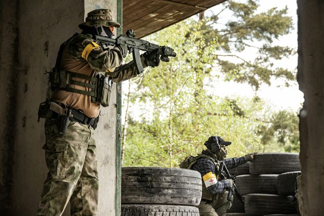 Joueur d'airsoft en position derrière un mur de pneus, coéquipier à l'abri à l'arrière-plan, sur un terrain aménagé