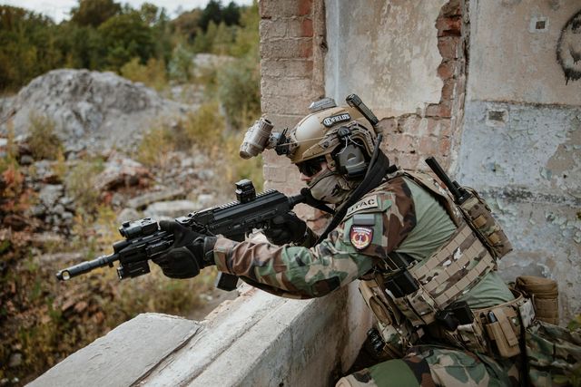 Joueur d'airsoft casqué en position derrière un muret en béton, réplique épaulée, dans un décor de ruines