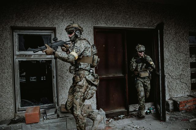 Deux joueurs d'airsoft en tenue camouflée progressent vers la porte d'un bâtiment abandonné, répliques en joue