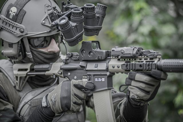 Joueur d'airsoft équipé d'un casque avec support de vision nocturne et casque audio, en joue avec une réplique