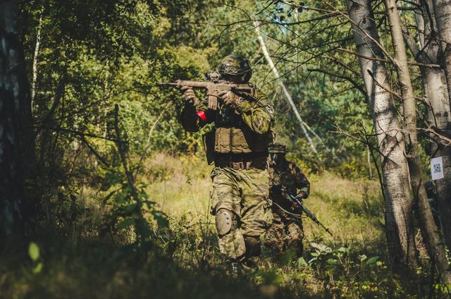 Joueur d'airsoft en tenue camouflage épaulant une réplique type M4 en sous-bois, un coéquipier en appui derrière lui